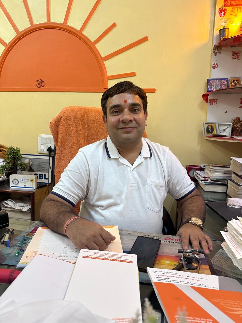 sitting at a desk with papers and a stethoscope in front of him, wearing a white shirt with a religious symbol on his forehead, and a background featuring a bright orange sun mural